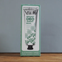 Load image into Gallery viewer, A boxed tube of Bergamot Deo Scented Balm standing upright on a wooden surface against a gray wall. The packaging is mint green and white with a printed tube illustration and text including 'BERGAMOT DEO SCENTED BALM' and 'SUSTAINABLY MADE FROM WOONY FRUITS.
