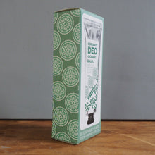 Load image into Gallery viewer, A tall rectangular box of DEO Ocean Balm standing upright on a wooden surface against a plain gray wall. The box is green and white with a repeating circular pattern on one side and product imagery on the front.
