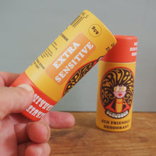 Load image into Gallery viewer, A hand holds a yellow and orange roll-on deodorant tube labeled 'Extra Sensitive' in front of another standing tube of the same brand on a wooden surface against a gray wall.
