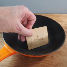 Load image into Gallery viewer, A hand holds a small rectangular wooden EcoLiving brand insert over a black frying pan with an orange handle on a wooden table.
