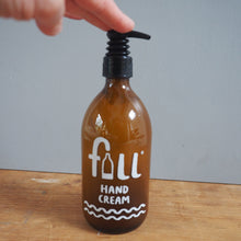 Load image into Gallery viewer, An amber glass hand cream bottle with a black pump dispenser, labeled 'fill hand cream' in white text, sitting on a wooden surface with a hand pressing the pump against a gray background.
