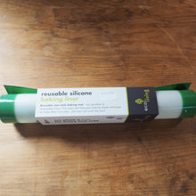Load image into Gallery viewer, A roll of reusable silicone baking liner by eco living, wrapped in a white, black, and green paper band, resting on a wooden tabletop.
