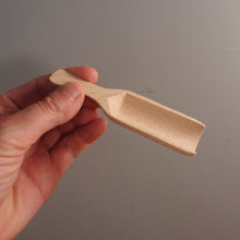 Load image into Gallery viewer, A hand holding a small, unfinished light wood scoop or miniature shovel against a plain gray background.
