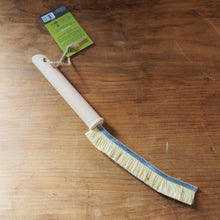 Load image into Gallery viewer, A small hand brush with a light wood handle and natural bristles, placed on a wooden tabletop with its green retail tag attached.
