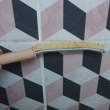 Load image into Gallery viewer, A hand holds a curved metal cleaning brush with natural bristles against a geometric tiled wall with pink, white, and black cubes.

